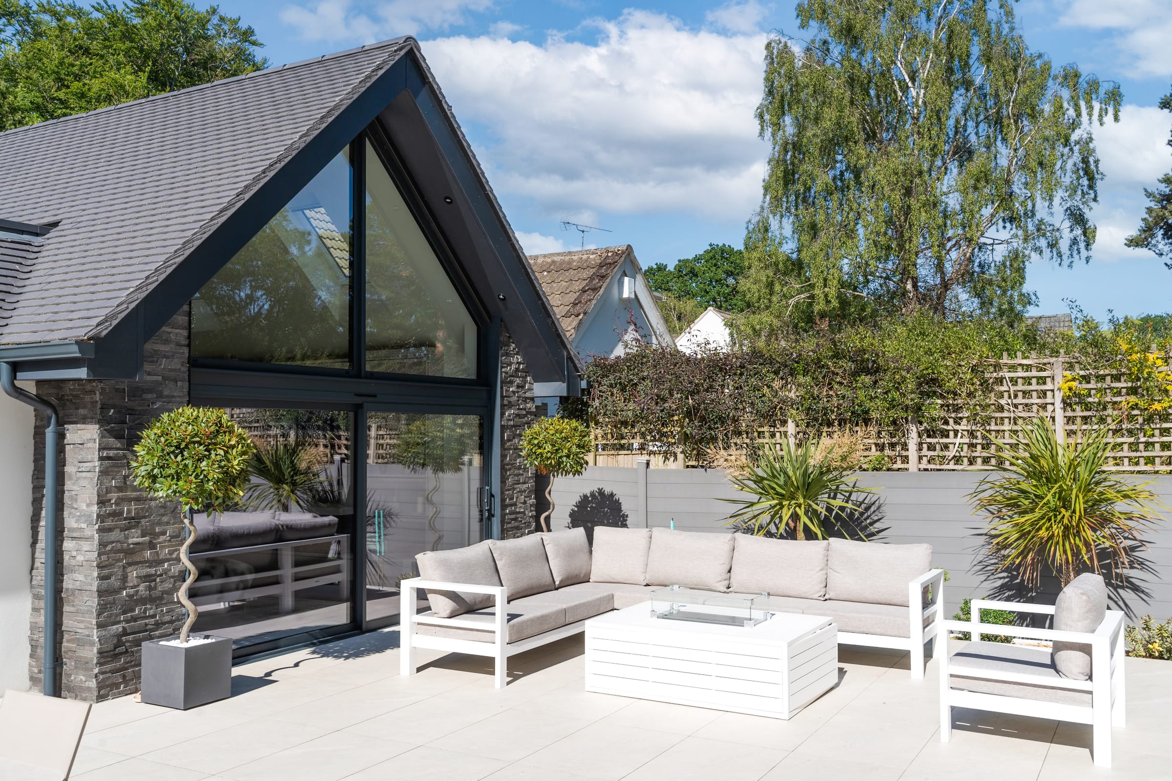 Anthracite aluminium sliding door under gable glazing, opening onto a paved terrace with garden furniture